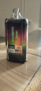 Dispositivo de vapeo DelphiTech negro con etiqueta holográfica "Yogur de fresa" y banderas europea y estadounidense, colocado sobre una superficie de madera.