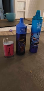 Three colorful vape devices are placed on a table: a small red and silver one labeled "Volt Cube," a blue one labeled "Bang Box" with a monster graphic, and a taller blue device labeled "Bang Beach" featuring another monster design.