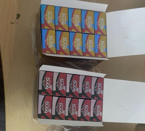 Due scatole aperte contenenti confezioni colorate di articoli a marchio "bang": la scatola superiore contiene confezioni gialle, blu e rosse, mentre la scatola inferiore contiene confezioni nere e rosa.